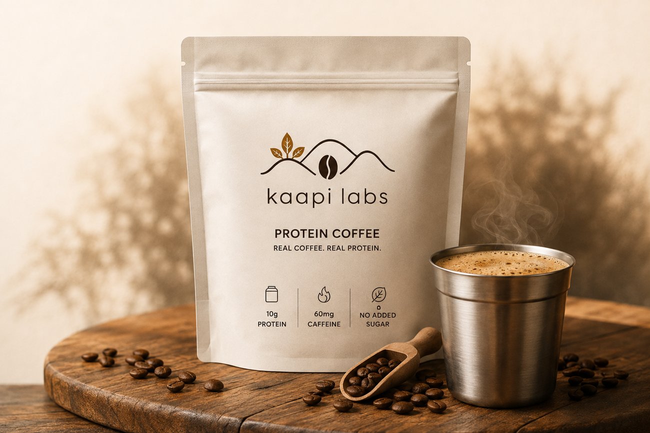 Kaapi Labs Protein Coffee pouch beside a steaming stainless steel tumbler of freshly brewed coffee, with wooden scoop and beans on a rustic wooden board